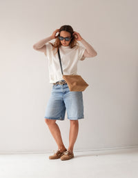 Mujer de pie luciendo un look casual de bermuda de jean y camisa blanca con mocasines apache color latte y bolso cruzado.