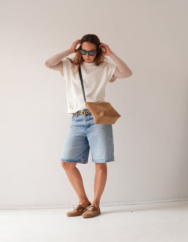 Mujer de pie luciendo un look casual de bermuda de jean y camisa blanca con mocasines apache color latte y bolso cruzado.