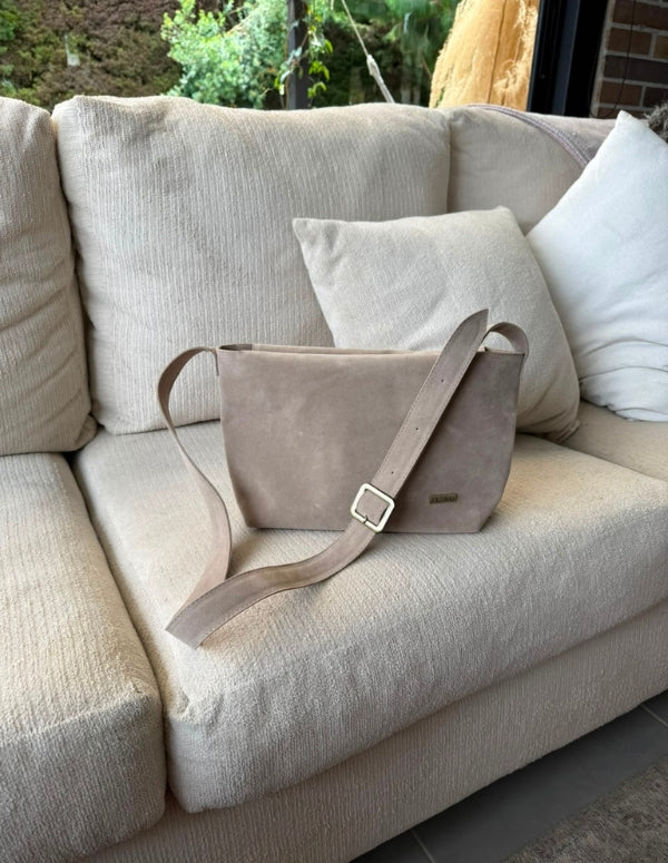 Bolso manos libres estilo bandolera color arena con correa ancha, descansando sobre un sofá claro. Diseño de marroquinería vegana.