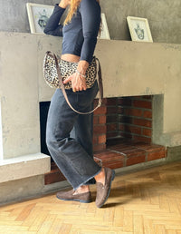 Imagen de estilo de vida que muestra a una persona de pie con vaqueros oscuros y mocasines marrones de gamuza Animalista cerca de una chimenea de ladrillo, sosteniendo un pequeño bolso con estampado de leopardo