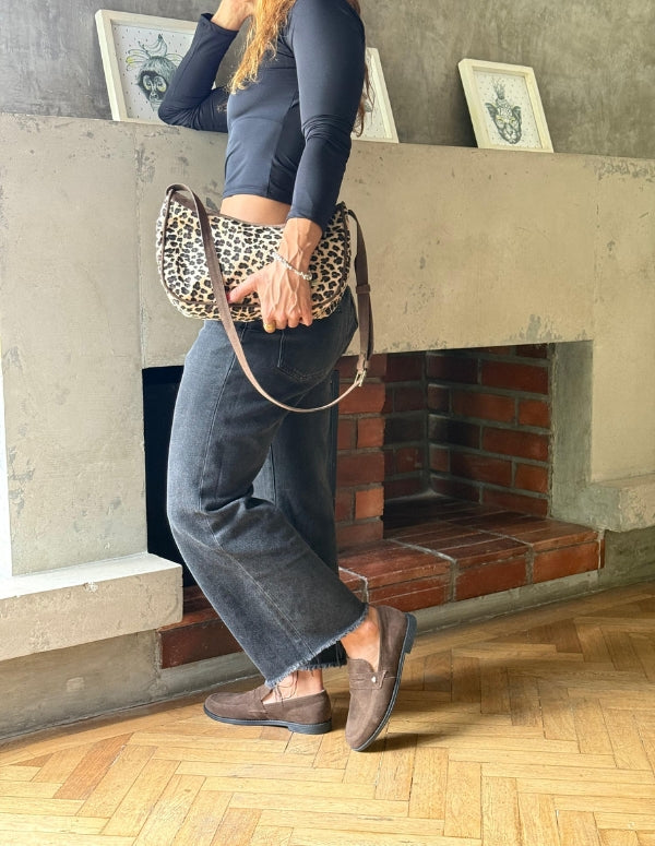 Imagen de estilo de vida que muestra a una persona de pie con vaqueros oscuros y mocasines marrones de gamuza Animalista cerca de una chimenea de ladrillo, sosteniendo un pequeño bolso con estampado de leopardo