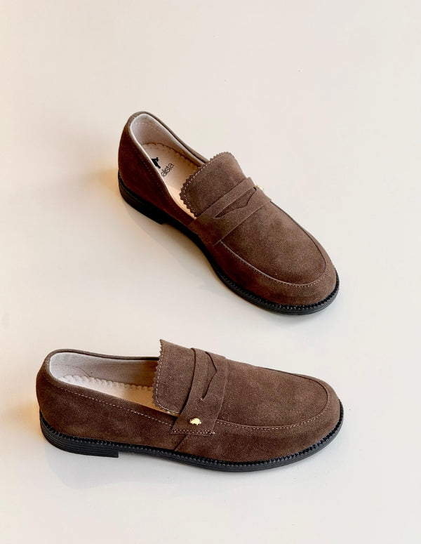 Primer plano superior de un par de mocasines tipo penny loafer de color marrón oscuro con textura de gamuza, de la marca Animalista, sobre un fondo blanco limpio