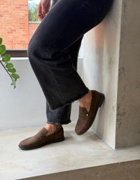Primer plano de estilo de vida de las piernas y el pie de una persona con pantalones oscuros, mostrando un mocasín marrón de gamuza Animalista.