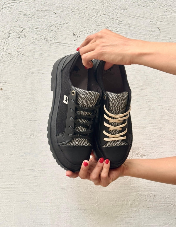 Manos sosteniendo un par de tenis negros para mujer de suela alta plataforma con detalles texturizados. Calzado urbano de moda consciente hecho en Colombia.