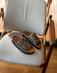 Par de sandalias cómodas tipo slide para mujer con banda ancha de estampado animal print de leopardo, descansando sobre una silla. Calzado vegano hecho en Colombia.