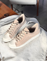 Primer plano detallado de tenis casuales blancos con panel lateral tejido en color beige. Calzado con cordones blancos, puntera de goma y suela plana sobre fondo de madera.