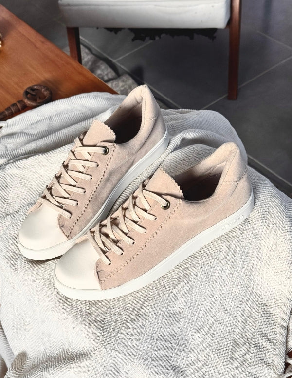 Primer plano detallado de tenis casuales blancos con panel lateral tejido en color beige. Calzado con cordones blancos, puntera de goma y suela plana sobre fondo de madera.