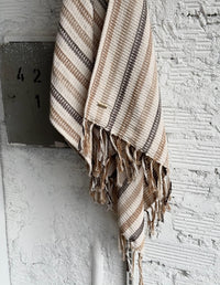 Poncho tejido para mujer en tono gris claro con terminación de flecos y marquilla metálica, colgado en una pared rústica blanca   . Ropa vegana Animalista.
