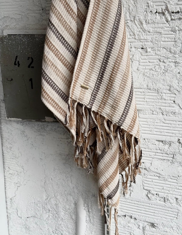 Poncho tejido para mujer en tono gris claro con terminación de flecos y marquilla metálica, colgado en una pared rústica blanca   . Ropa vegana Animalista.