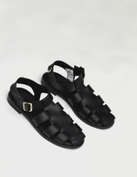 Par de sandalias planas color negro para mujer, diseño estilo cangrejera con tiras cruzadas y ajuste de hebilla en el tobillo. Calzado vegano hecho en Colombia.
