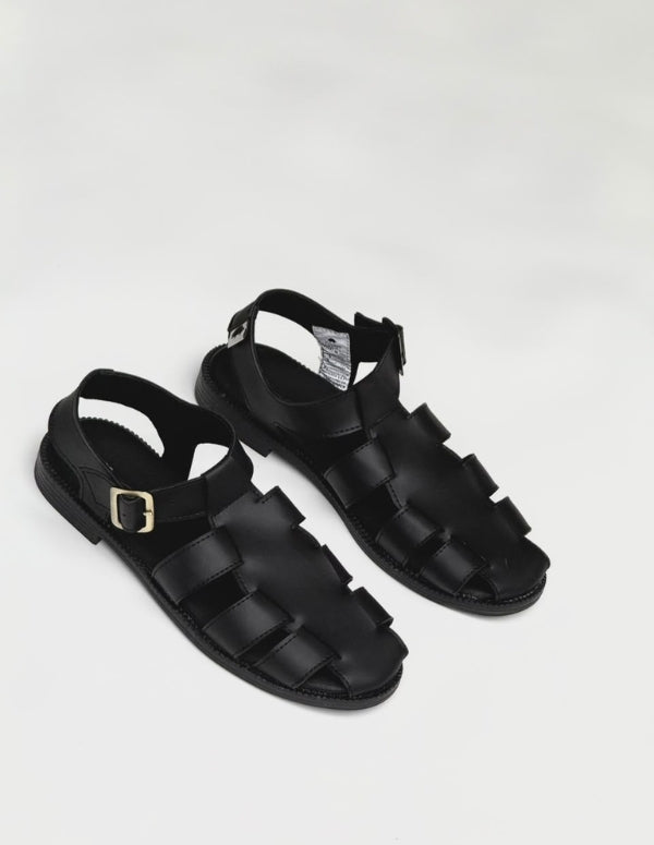 Par de sandalias planas color negro para mujer, diseño estilo cangrejera con tiras cruzadas y ajuste de hebilla en el tobillo. Calzado vegano hecho en Colombia.