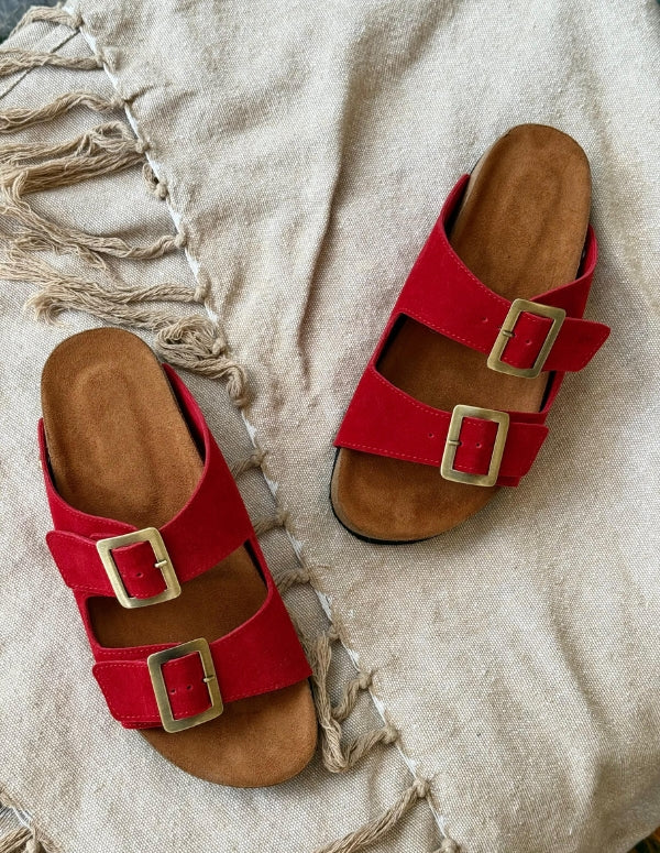 Par de sandalias rojas para mujer con diseño de dos correas y hebillas metálicas sobre fondo de tela cruda. Calzado cómodo vegano hecho en Colombia.