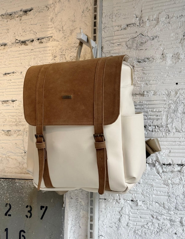 Morral de trabajo para mujer con cuerpo beige y tapa color latte (café claro), colgado en una pared de ladrillo blanco texturizado. Diseño de moda ética.